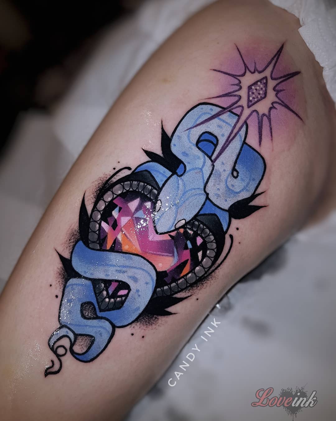 a tattoo of a blue snake wrapping around a heartshaped jewel. the snake and jewel have highlights of pink and purple. there is a pink and purple sparkle emanating from a smaller jewel above the heart. the tattoo is outlined in black and white and sits on someone’s upper thigh.