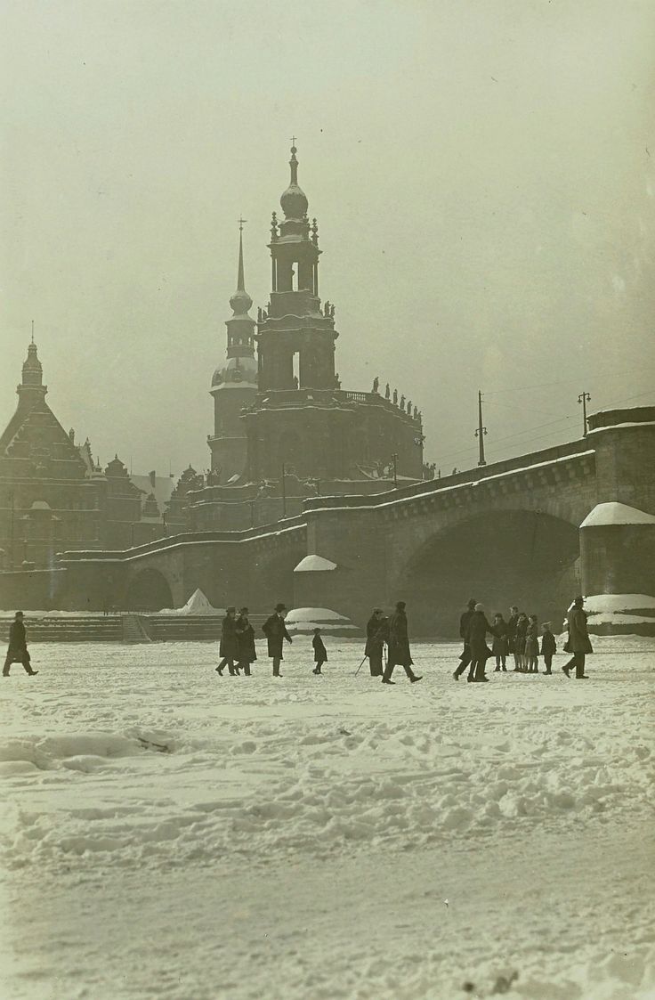 a black and white vintage photograph of dresden, germany in winter. the frauenkirche is prominently featured in the background, with a bridge spanning the elbe river in the foreground.  the river is frozen over, and several people are walking across the snowcovered surface. the overall atmosphere is cold and historical.  the image provides a glimpse into daily life in dresden during the early 20th century.