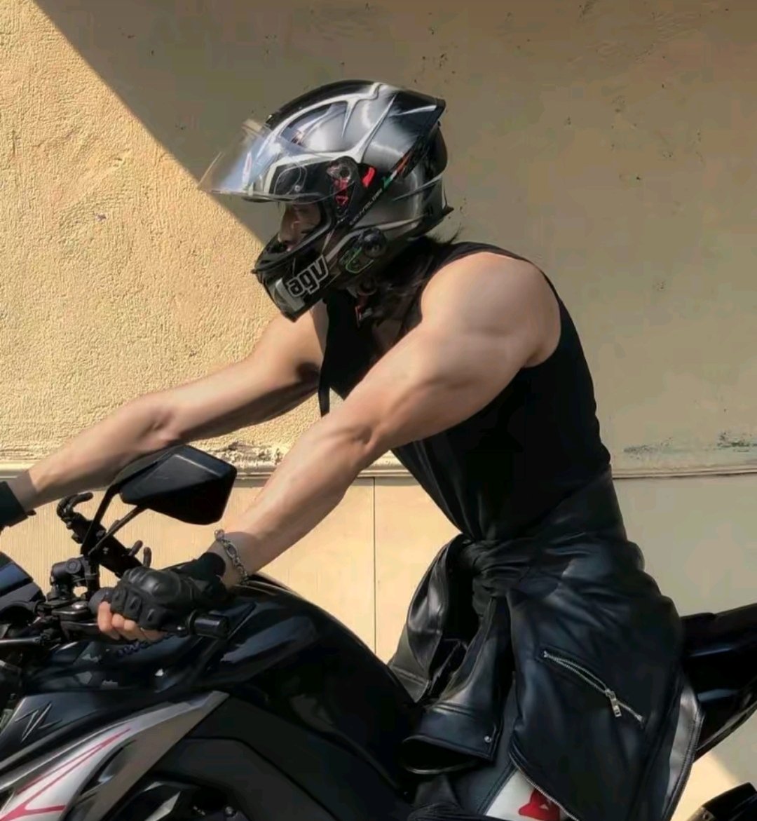 a man wearing a black leather jacket and a motorcycle helmet, sitting on a black motorcycle, looking to the side. the man is muscular and handsome, and the photo captures a sense of cool, speed and freedom. the setting is outdoors, possibly in a city.