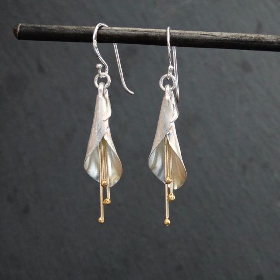 this is a closeup shot of a pair of silver fuchsia flower earrings. the earrings have a sleek and modern design, with a long, slender stem and a delicate flower at the top. the earrings are made of sterling silver and are accented with gold. the background is a dark gray color, which helps to highlight the beauty of the earrings.
