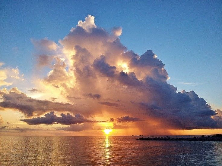 the image showcases a breathtaking sunset over a calm ocean.  a massive, dramatic cumulonimbus cloud dominates the sky, its base illuminated with vibrant oranges and yellows from the setting sun, while its upper portion is a darker grey. rain appears to be falling from a portion of the cloud. the sun is partially visible, setting below the horizon line. the ocean reflects the warm colors of the sunset. a dark shoreline or jetty is visible in the lowerright portion of the image. the overall mood is one of aweinspiring natural beauty and dramatic weather.