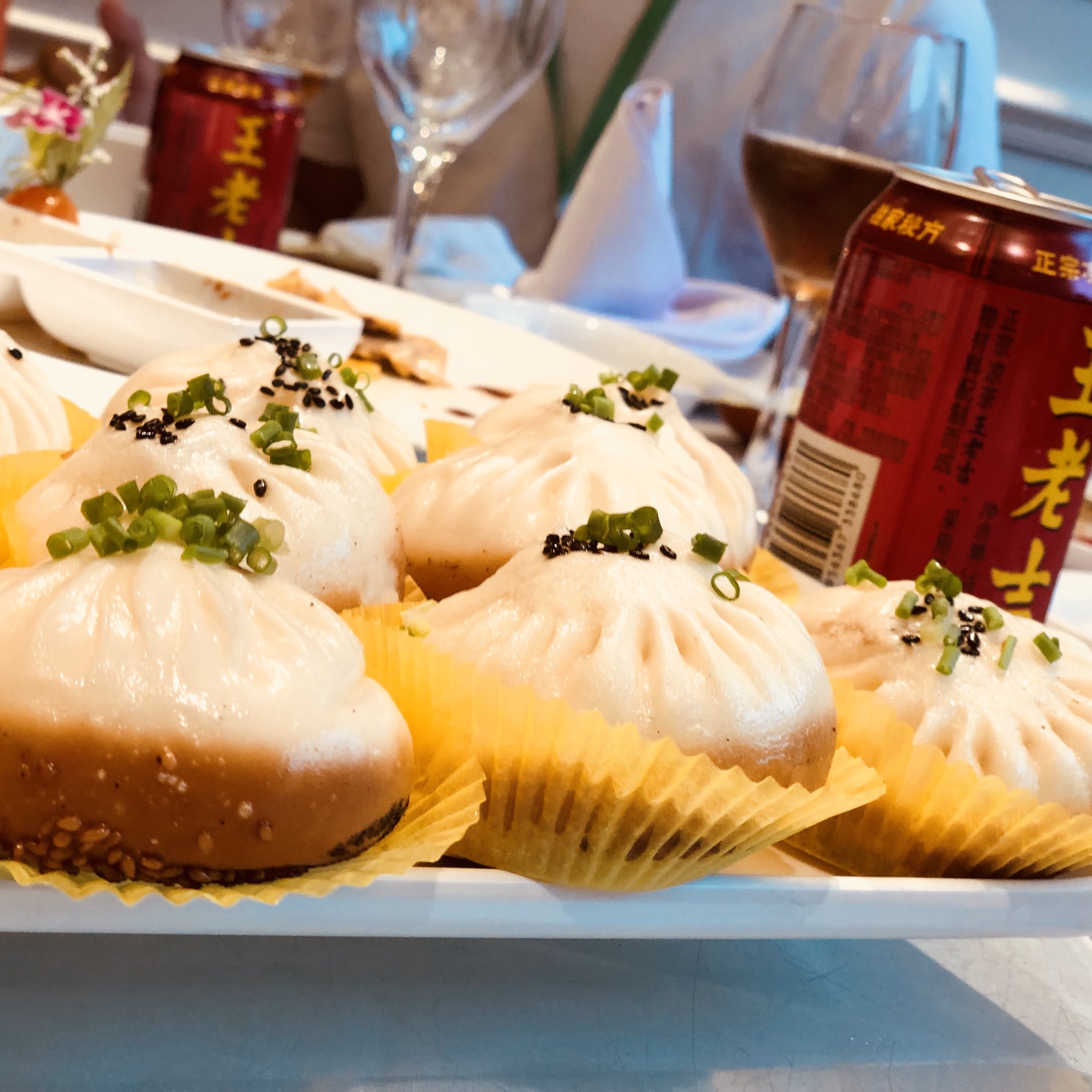 closeup photo of a plate of delicious steamed buns (baozi) on a table set for a meal. the buns are garnished with green onions and black sesame seeds.  there are cans of beer, wine glasses, and other plates in the background.