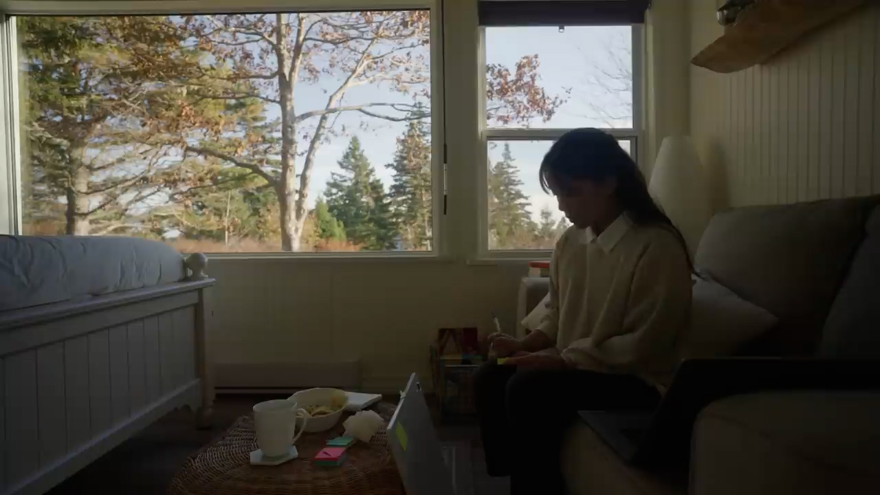 he image shows a young woman sitting on a couch in a bedroom, studying. she is wearing a white sweater and black pants. she is looking down at a book and appears to be focused on her work. on the floor in front of her is a laptop, a cup, a plate, and some other items. there is a bed in the background and a window to the left of the woman. the window is large and bright, and it looks out onto a forest. the room is lit by natural light coming through the window, creating a warm and inviting atmosphere. the overall tone of the image is calm and peaceful.