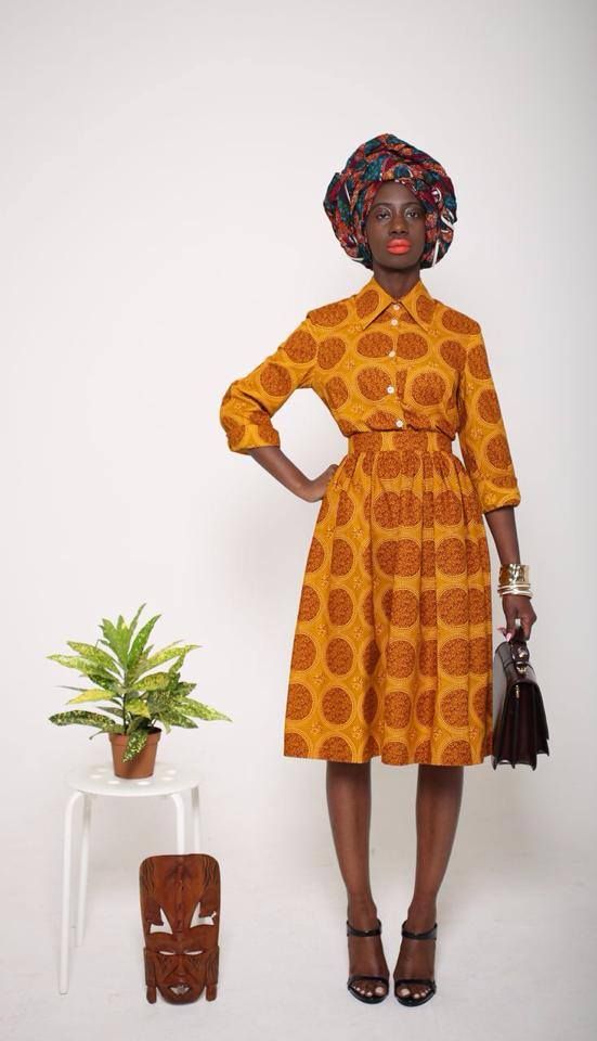a stylish darkskinned woman is standing against a white background. she's wearing a stunning mustard yellow african print midi dress with a high waisted pleated skirt and matching threequarter length sleeves. her outfit is complemented by a vibrant headwrap. a black handbag and black highheeled sandals complete her look.  a small plant sits on a white stool next to a carved wooden mask.  the overall aesthetic is one of sophistication and cultural pride.
