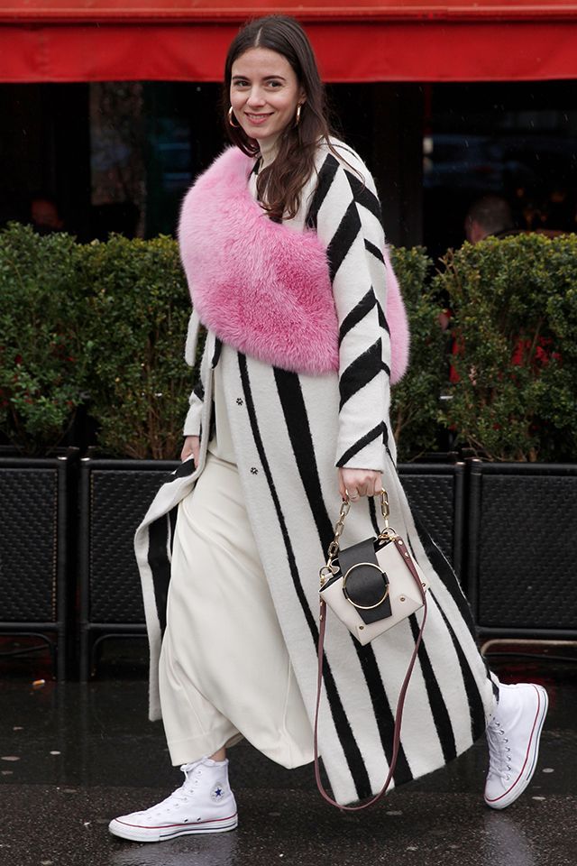 the image shows a stylish woman walking down a parisian street. she's wearing a long black and white striped coat over a creamcolored pant or jumpsuit.  a standout detail is the bright pink fur stole she's wearing.  she's carrying a stylish offwhite and black handbag and sporting classic white converse hightop sneakers. the overall aesthetic is chic and effortless street style. the background features greenery and the red awning of a building, suggesting a parisian setting.