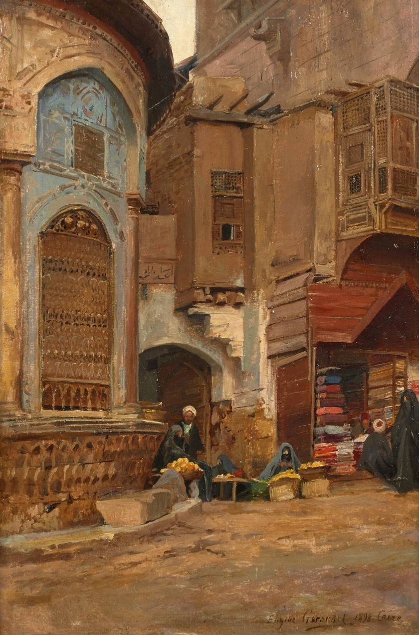 the painting depicts a busy street scene in an orientalist setting, likely a marketplace. several buildings with unique architectural details line the street, including a tall structure with a blue facade and ornate wooden window coverings. vendors are set up along the street selling goods. a man in a turban is sitting with baskets of fruit, possibly oranges. several women in veils are present, some appearing to be shopping. an awning shades a storefront displaying stacks of colorful textiles. the scene is rendered in warm, earthy tones with visible brushstrokes, characteristic of an oil painting.