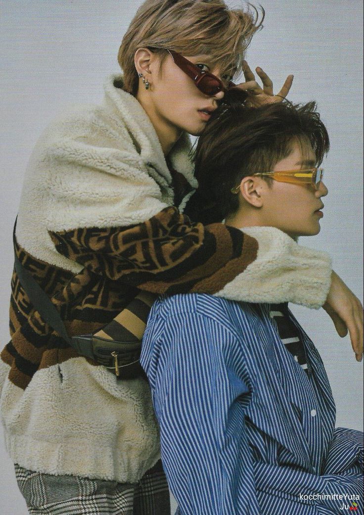 two young men in a studio shot with a white background. one man is wearing a white fuzzy jacket with a brown and black pattern, and the other is wearing a blue and white striped shirt. the man in the jacket has one arm around the other man and is leaning his head into his shoulder. the man in the striped shirt is sitting and looking away from the camera. both men are wearing sunglasses. the man in the jacket has brown hair and the other man has black hair. they are both smiling. the photo has a cool and stylish vibe.