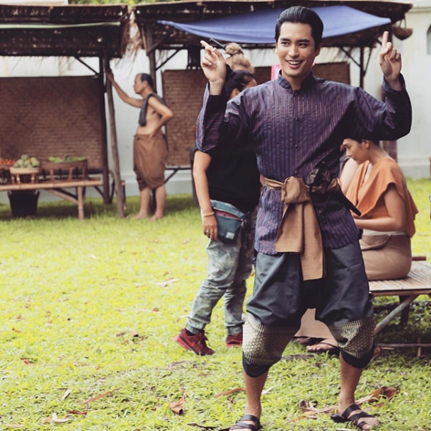a man in traditional thai clothing poses playfully on a grassy field, surrounded by other people. he's wearing a purple striped shirt, loose dark pants, and a brown sash. to his left, another man in jeans and a black shirt is partially obscured. in the background, an older man in a brown wrap stands near market stalls set up under a wooden structure.  two women are seated on a bench behind the posing man. the lighting suggests an overcast day, casting soft shadows.