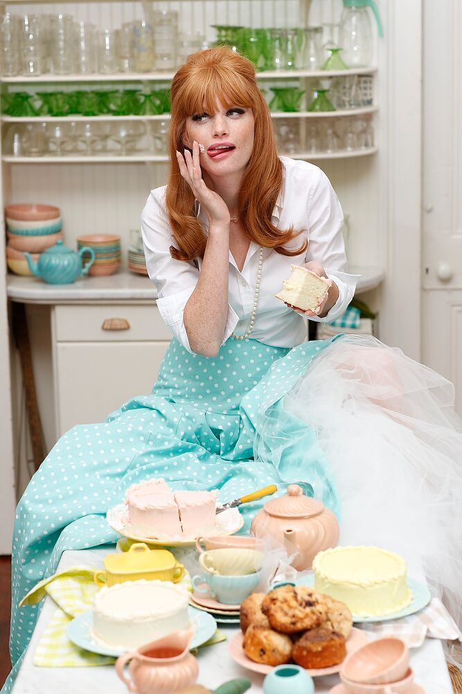 a playful redhead in a retro kitchen indulges in a variety of cakes. she's dressed in a blue polka dot skirt and white blouse, her expression a mixture of mischief and delight.  the scene is filled with pastelcolored cakes, teacups, and vintage kitchenware, creating a playful and nostalgic atmosphere.