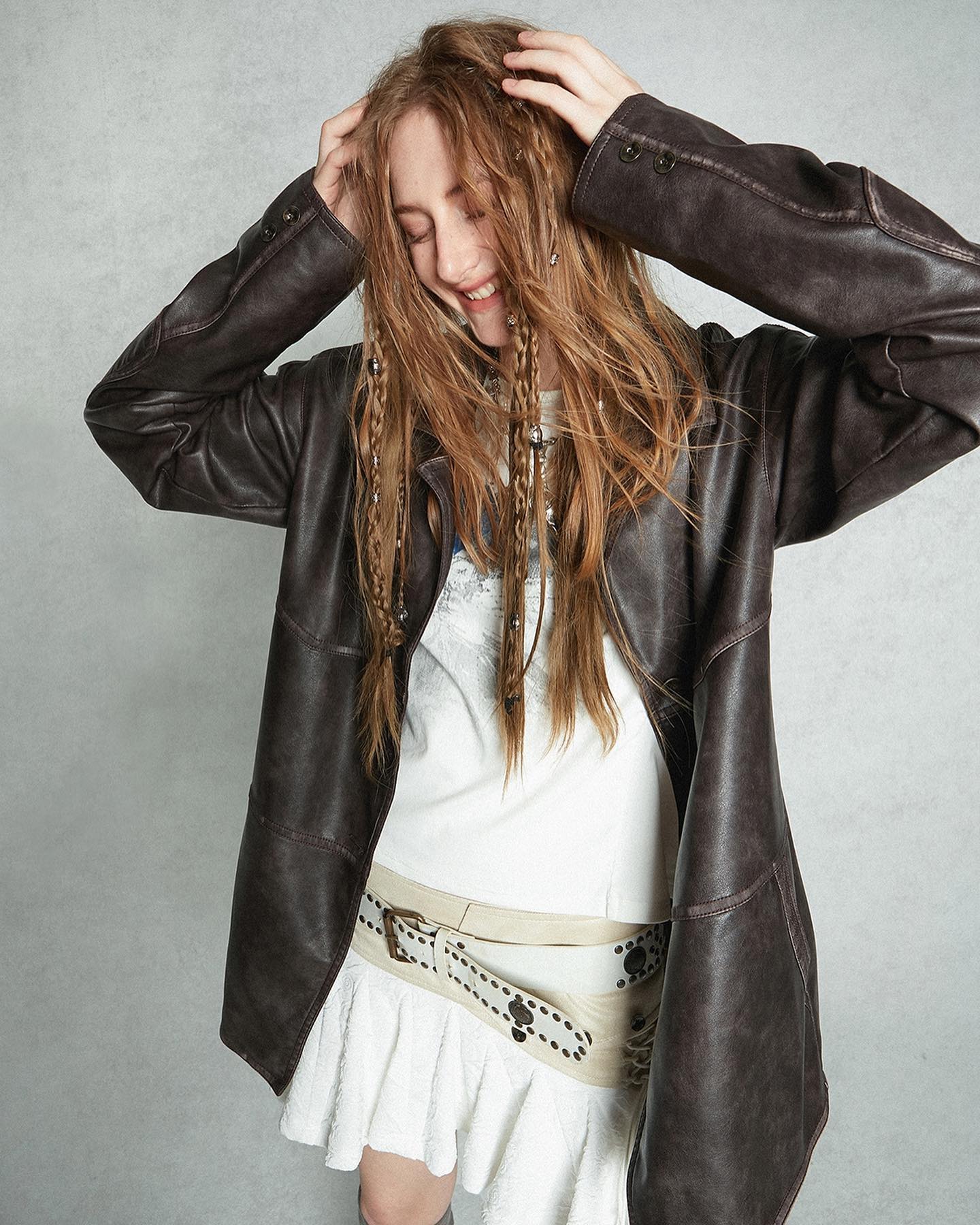 a young woman with long, braided hair is posing in a studio shot. she is wearing a brown faux leather jacket, a white shirt, a white ruffled skirt and a beige studded belt. her hair is styled in loose braids adorned with beads. she is smiling and touching her hair. the background is a neutral gray. the overall style is casual, trendy and slightly bohemian.