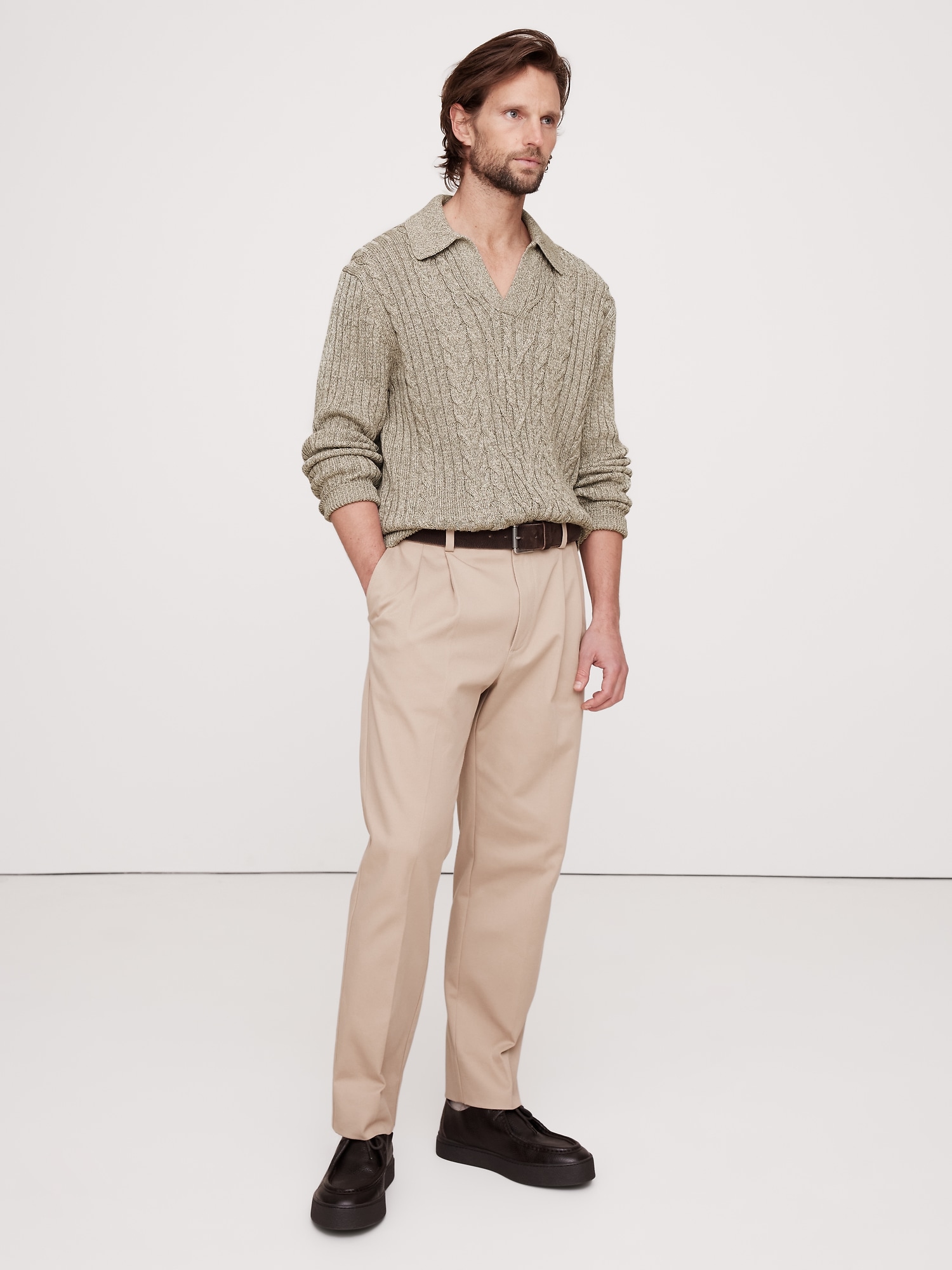 a man stands in a full shot, facing slightly right. he wears a sage green cable knit sweater with a collar, khaki pants with a brown belt, and black shoes. the background is a plain, light studio setting.