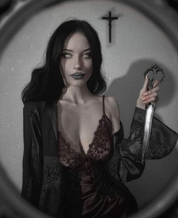 a young woman with long dark hair is looking directly at the camera while holding a sharp knife. she is wearing a black robe and red lace lingerie. she is surrounded by dark shadows with a cross in the background. the photo has a dark and gothic feel to it, suggesting danger and mystery.
