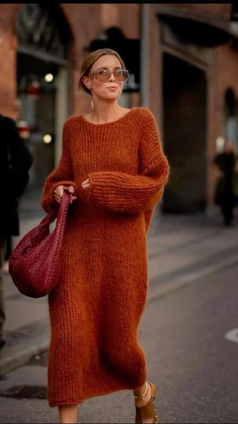 a woman in an orange knitted dress and a brown bag is posing on the sidewalk. she is wearing sunglasses and has long brown hair. the image is taken from a street perspective and captures the woman from the waist up. the background is blurred and features an urban environment with buildings and trees.