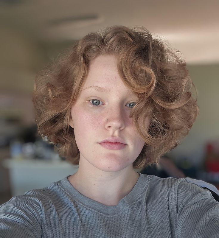 a closeup portrait of a woman with curly red hair, wearing a gray shirt. she has a nose ring and is looking directly at the camera.