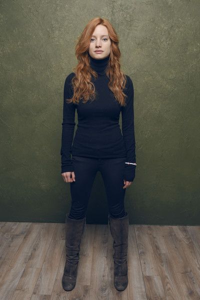 a woman with long red hair stands in front of a green background. she is wearing a black turtleneck sweater, black pants, and brown boots. she has a silver bracelet on her right wrist. the image is lit with soft lighting.