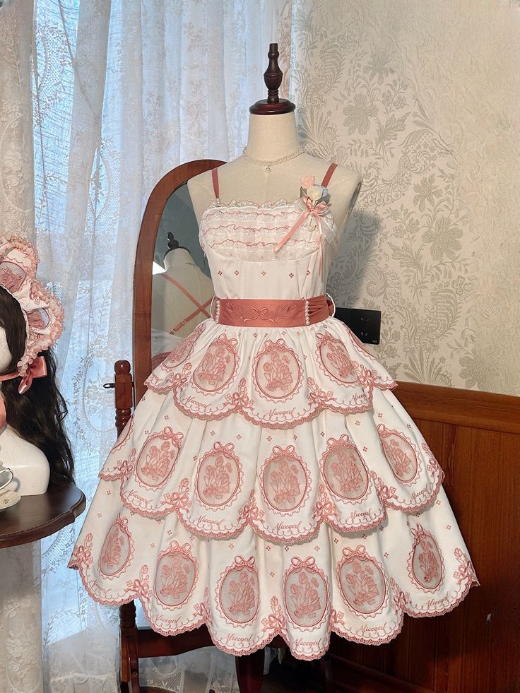 a white and pink lolita fashion dress is displayed on a mannequin. the dress is short with a tiered skirt and delicate embroidery. a matching headpiece is on a stand in the background. the room is decorated in a vintage style.