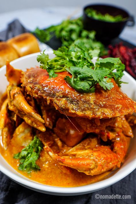 a closeup shot of a bowl of chili crab, a signature singaporean dish. the crab is cooked in a rich, orangered chili sauce and is garnished with fresh coriander.  the bowl sits on a dark surface, with steamed buns (mantou) and some additional cilantro visible in the background. the image is professionally styled, emphasizing the vibrant colors and textures of the food. the watermark "nomadette.com" is visible in the lower right corner.