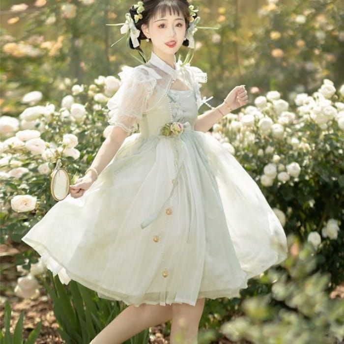 a young woman in a flowing, pale green dress stands in a garden. the dress has sheer sleeves, a high collar, and is decorated with floral embroidery. she is posing with her hand on her hip and a gentle smile on her face. the image has a soft, dreamy quality.
