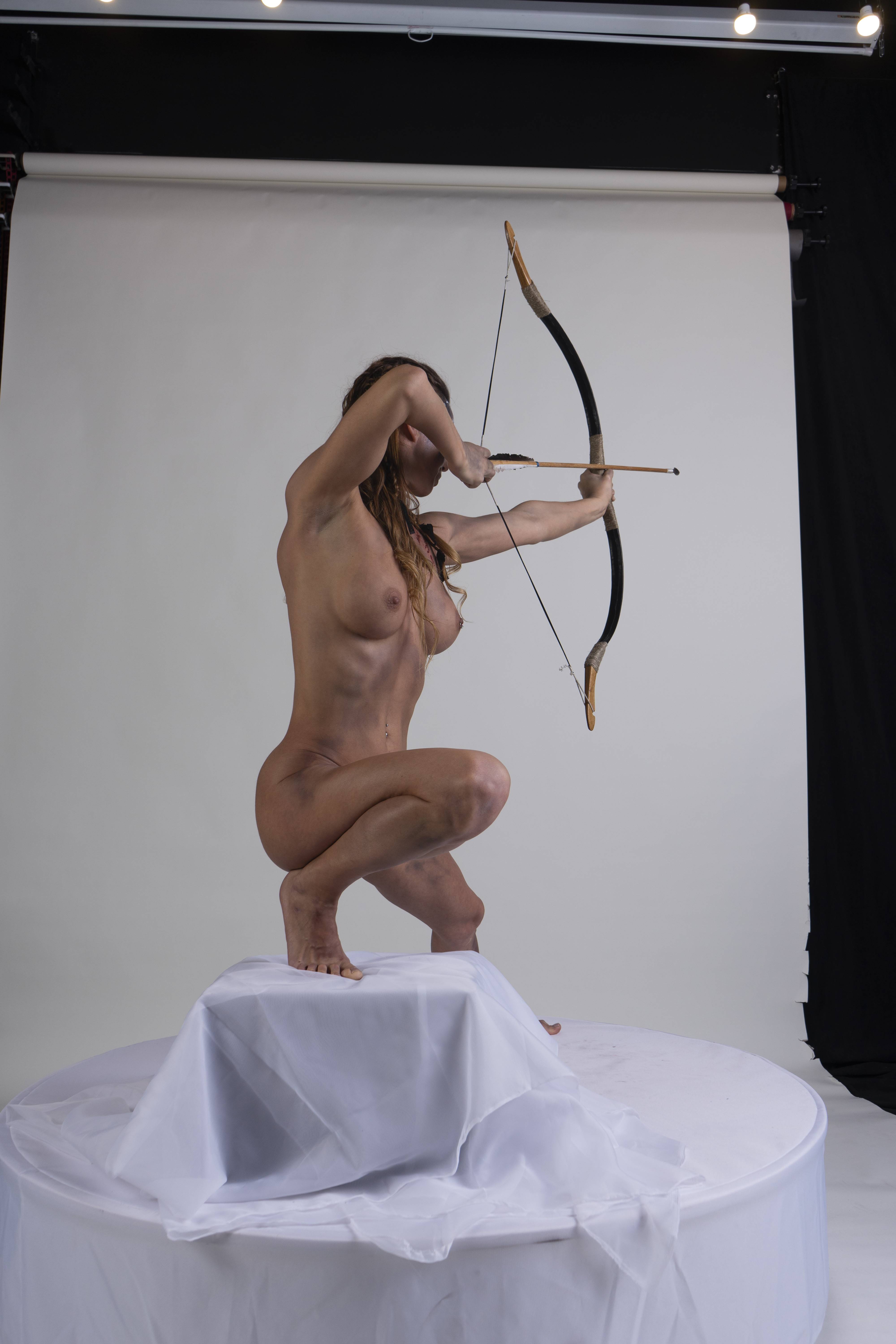 a nude woman is squatting on a platform draped with white fabric, holding a bow and arrow. she is aiming towards the right of the frame. the background is a studio setting with a white backdrop and black curtains on the right.