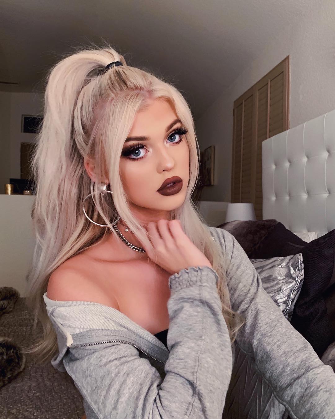 a young woman, likely loren gray, with platinum blonde hair styled in a high ponytail, stands in what appears to be a bedroom. she is wearing large hoop earrings, dark lipstick, and a gray sweatshirt that is partially offtheshoulder. she also has on a chain necklace. her hair is pulled up in a high ponytail with strands framing her face. she has blue eyes and appears to be wearing full makeup. she is resting her hand near her neck. behind her is a bed with pillows, a white headboard, and what appears to be closet doors in the background.