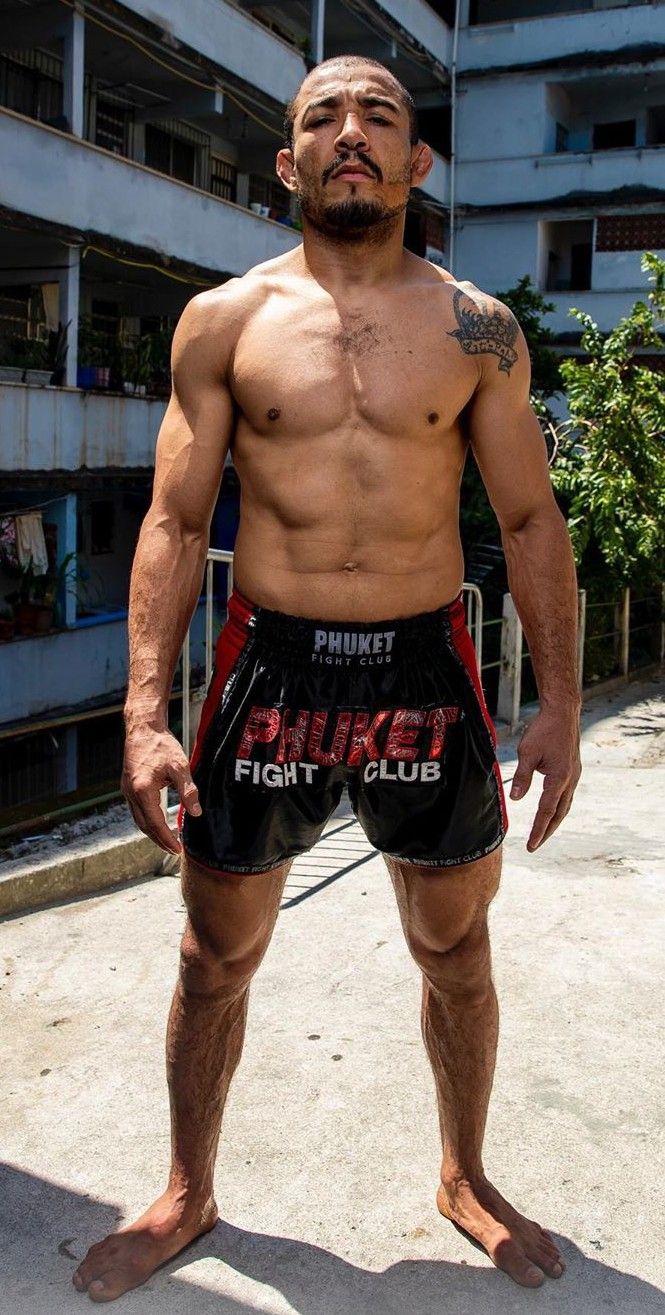 a shirtless man wearing fighting shorts stands with his arms at his side. the man is muscular and has a tattoo on his upper left shoulder. he is looking at the camera. the backdrop is an outdoor urban environment. the shorts say "phuket fight club" on them.
