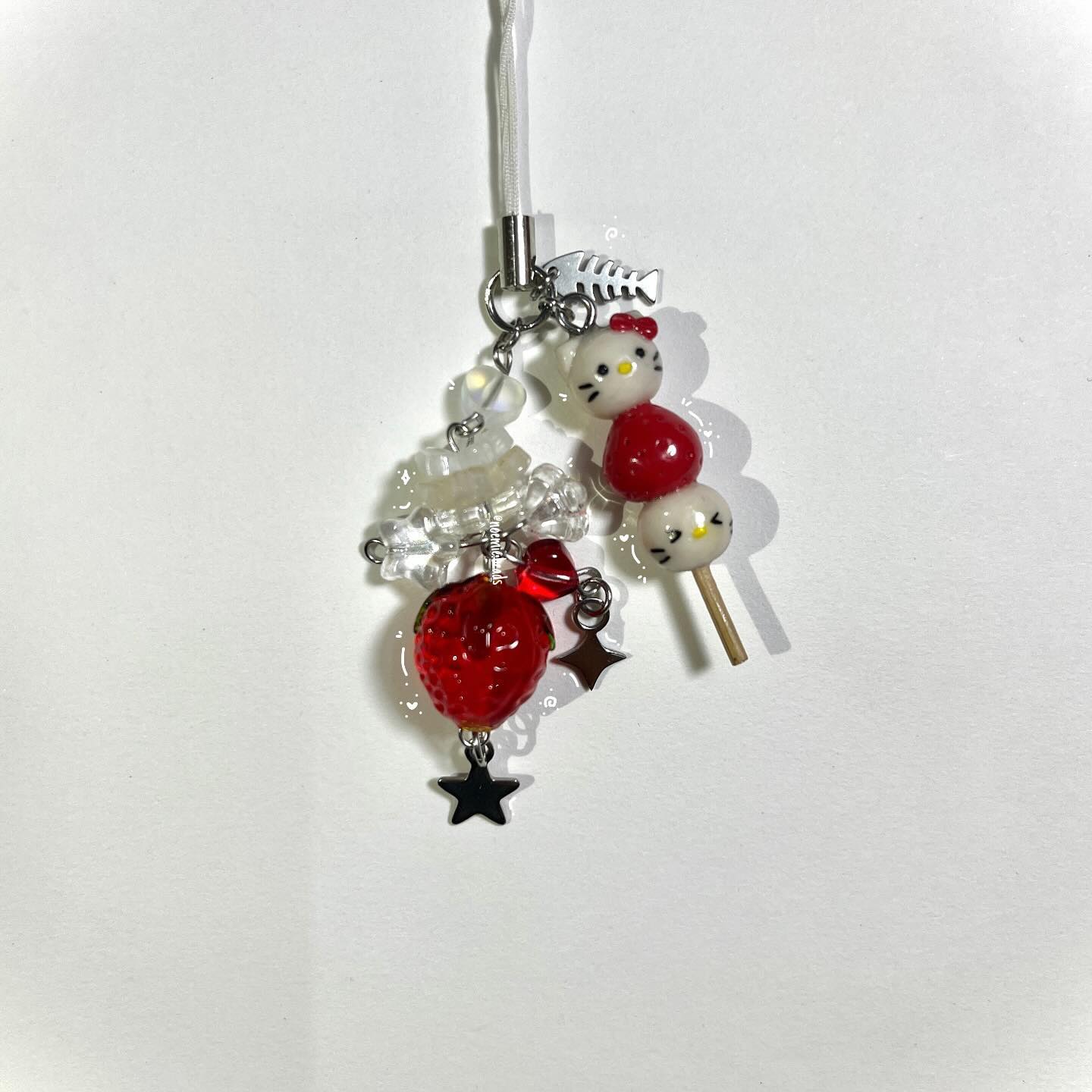 a charm made with beads and other objects. it consists of a clear cord at the top, a fish charm, clear and white beads, a red strawberry charm, star charms, and a dango stick charm made of a hello kitty head, a red bead, and a face bead.