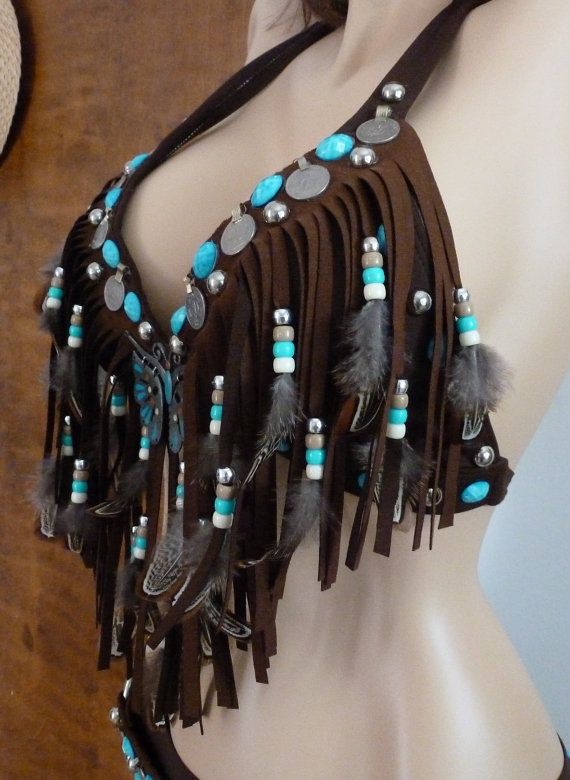 a closeup of a brown leather bra with turquoise beads, feathers, and fringe. it is a handmade piece of jewelry that would be perfect for a bohemian or festival style. the bra is accessorized with many other jewelry pieces.