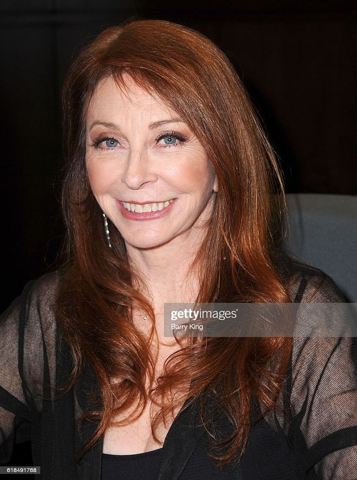 a close up portrait of a woman with long red hair smiling at the camera. she's wearing a black sheer top with a lace design. she is sitting on a black chair in a dimly lit room.