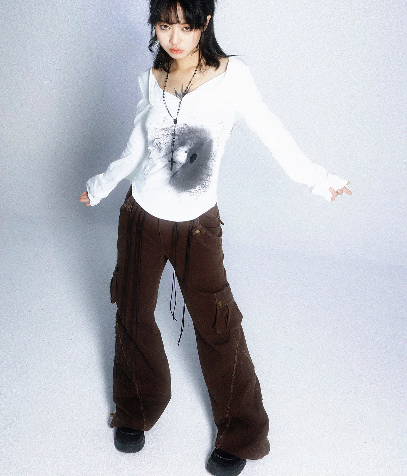 young asian woman with dark hair is posing in a studio shot against a white background. she is wearing a white longsleeved shirt with a dark graphic print, dark brown wideleg cargo pants, and black boots. her shirt is low vneck and she is wearing a black cross necklace. her overall style is grunge, alternative, and a bit dark academia. the image is welllit and the model is clearly visible.