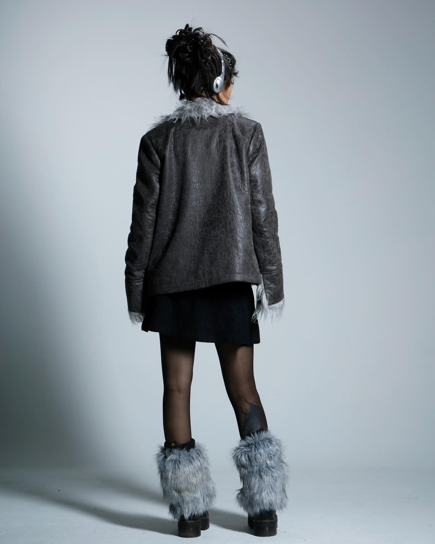 a full shot of a woman from the back, wearing a dark grey jacket with a fur trim, a black skirt, black tights, and grey fur leg warmers. she has headphones on and her hair is in a bun. the background is a plain light grey.