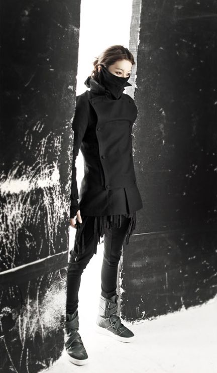 a young woman wearing a black coat and boots stands in a doorway. she is wearing a black face mask.  the woman is looking down at her feet. the photo is taken in a black and white style and creates a cool and stylish image.