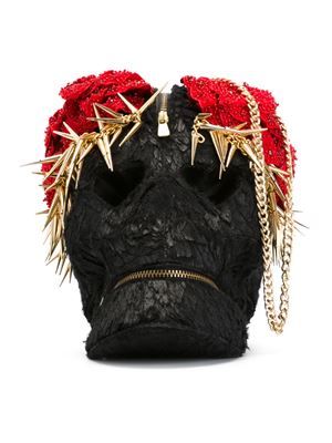 the image shows a black skullshaped purse with a red flower decoration on top, adorned with gold spikes and a gold chain. the purse has a textured surface and features zipper closures.