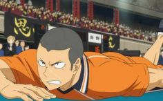 an anime character with short dark hair wearing an orange and black uniform is shown diving on a court. the character is drawn in an intense moment, eyes focused on an unseen object. he is surrounded by blurred figures in the background, suggesting a large crowd. banners and a scoreboard are visible in the background.