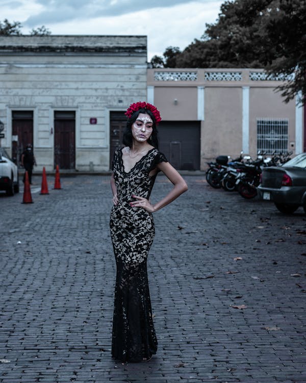 the image shows a woman dressed as a catrina, a figure associated with the day of the dead celebration in mexico. she is wearing a black lace gown and a red flower crown. her face is painted with the characteristic calavera catrina skull makeup. she stands on a cobblestone street in front of old, colonialstyle buildings.  motorcycles are parked in the background, and the overall atmosphere is moody and vintage. the photograph has a style that evokes a sense of tradition and cultural celebration.