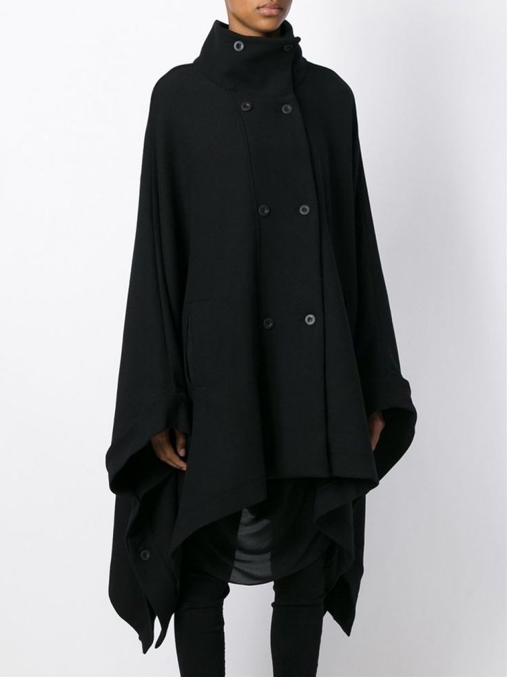 a person stands in front of a white background wearing a black cape. the cape features a high neckline with a button fastening, an asymmetrical hem, and a button fastening on the front. they are wearing leggings and dark under garments. the cape is worn as outerwear, and the overall style suggests layering. the cape is a ponchostyle garment.