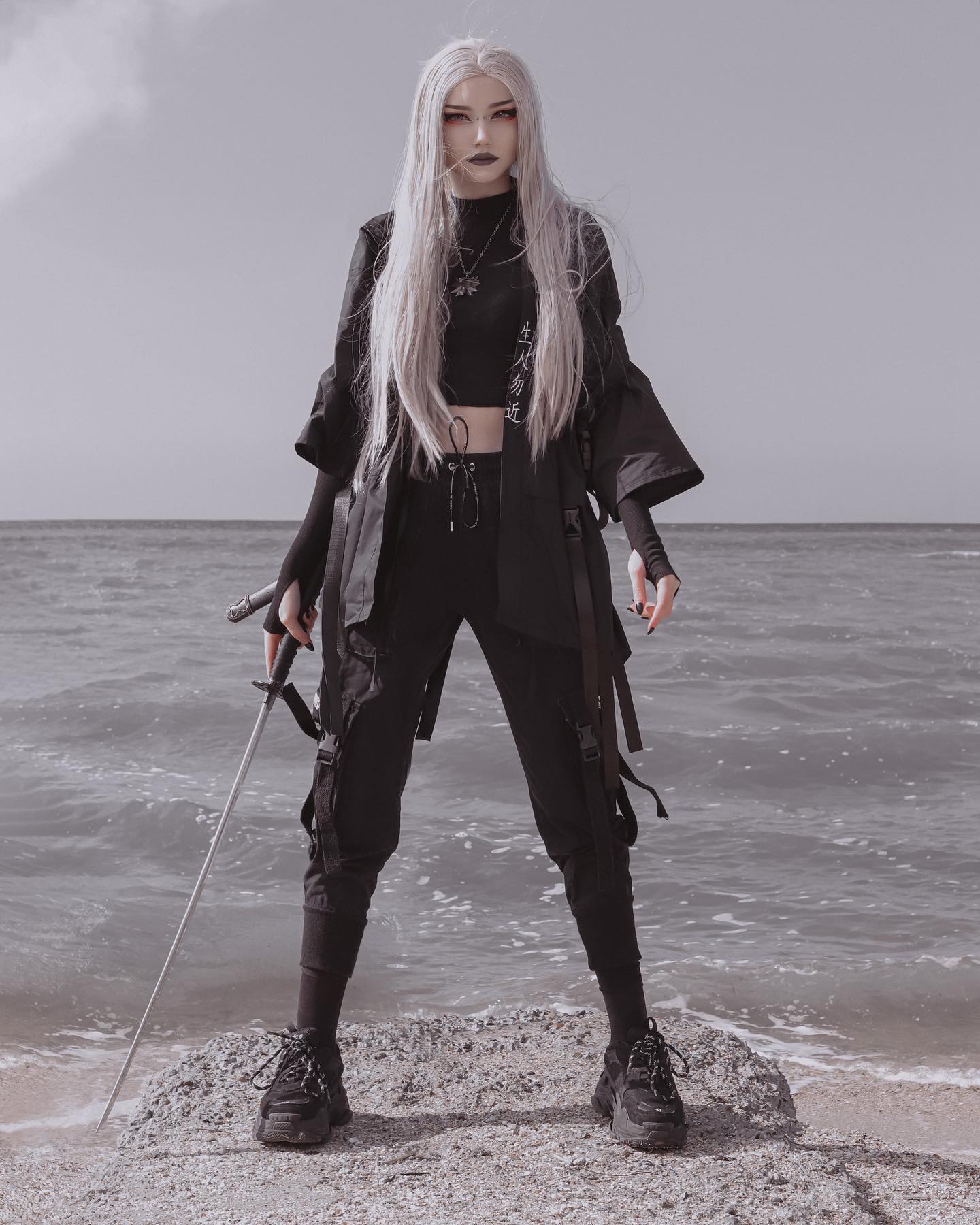 the image showcases a young woman with long, platinum blonde hair styled straight. she's dressed in a dark, edgy outfit: a black kimonostyle jacket over a black crop top and black cargo pants. the pants feature functional buckles and straps. she's holding a long, thin, silver sword, adding to the dramatic, almost warriorlike aesthetic.  her expression is serious and intense. the background is a moody, greytoned ocean and beach setting. the overall atmosphere is dark, stylish, and mysterious, with a blend of gothic, cyberpunk, and possibly animeinspired elements in the fashion. the woman stands on a rock near the water's edge.