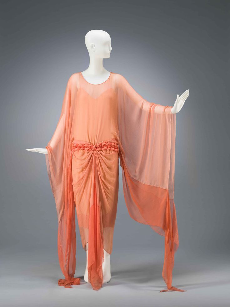 a peachcolored chiffon dress with long, flowing sleeves is displayed on a white mannequin. the dress features a rosette detail at the waist and draping throughout. the background is a neutral gray.