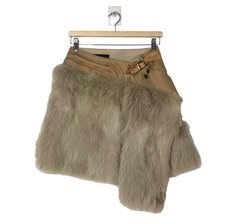 a beige fur skirt with brown leather accents, hanging on a wooden hanger. the skirt features a buckle detail at the waist and a unique asymmetrical design. it appears to be a highfashion piece that exudes elegance and sophistication.