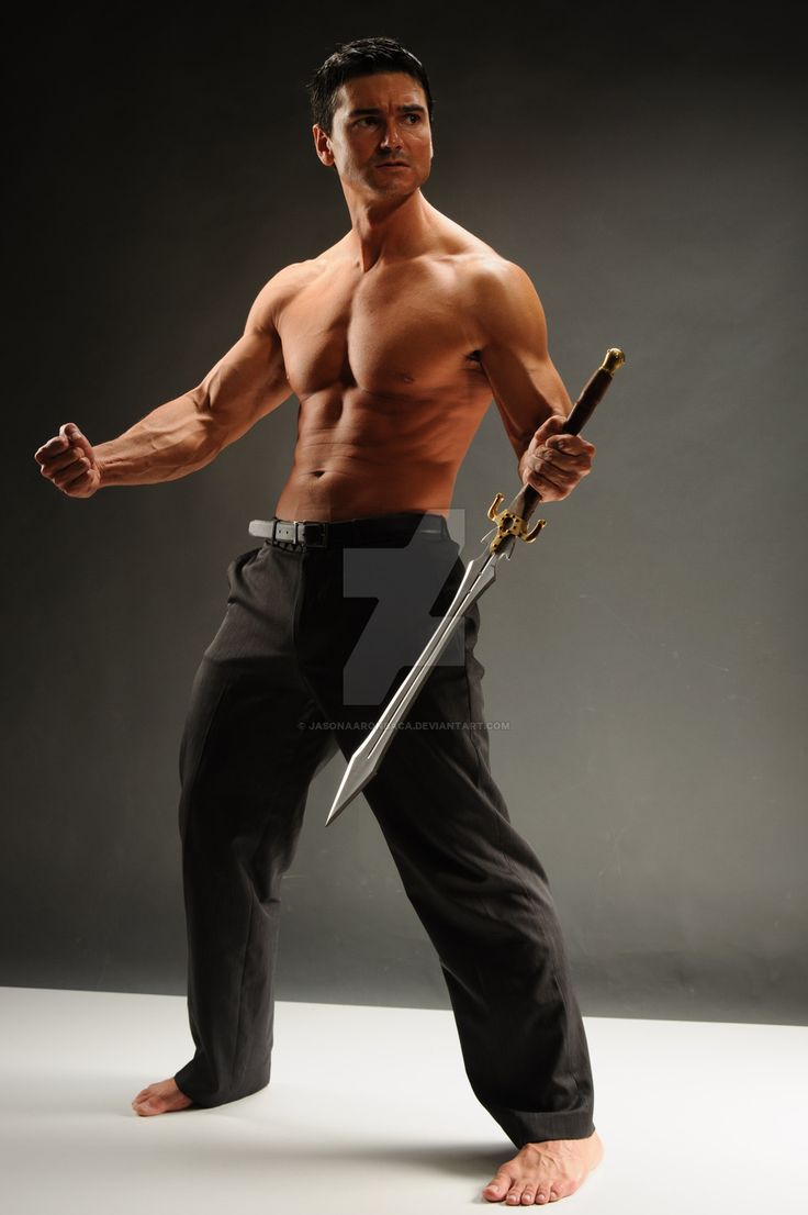 a shirtless man is holding a sword in his right hand. he is standing with his left leg forward and his right leg back. the man is muscular and fit. he has a serious expression on his face. the background is a simple grey.