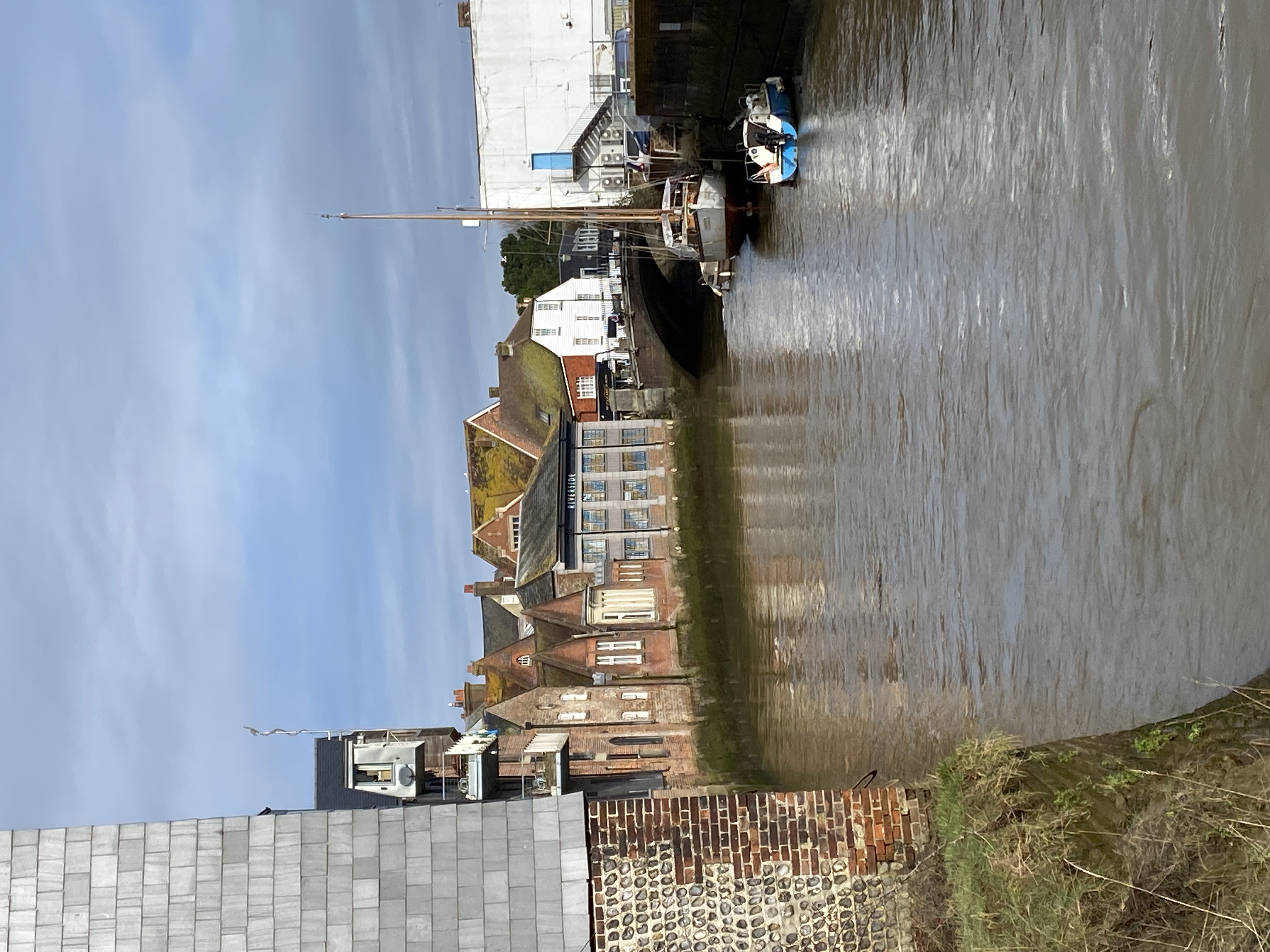 a picturesque view of a river with boats docked on the water. the river flows through the center of the image, flanked by brick and stone buildings on either side. the houses are mostly three stories tall and feature a variety of architectural styles.  a bridge crosses the river in the background, connecting the two sides. the sky is a bright blue with wispy white clouds. the scene is captured during the day, bathed in the warm light of the sun. the image evokes a sense of peace and tranquility, with a hint of history and charm.