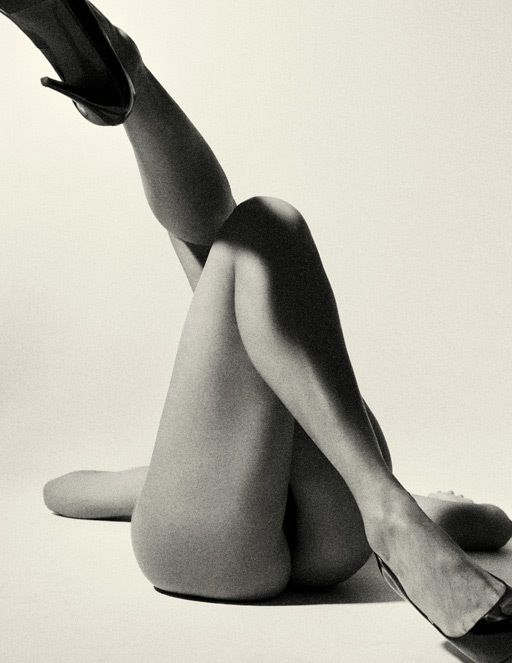 the image is a black and white, closeup photograph of a woman's body.  only her legs and torso are prominently displayed; the rest of her body is partially visible. she is lying on her side, her legs intertwined and angled, one leg extending upward with a highheeled shoe prominently displayed. the lighting is dramatic, casting strong shadows and highlights on her skin, enhancing the curves and texture. the overall mood is sensual and artistic, with an emphasis on the female form and the interplay of light and shadow. the image suggests a fashion or fine art photography style.