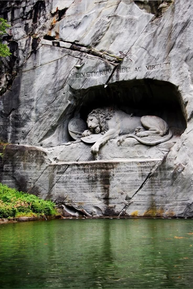 the image shows the lion monument in lucerne, switzerland. the monument is a rock relief depicting a dying lion, carved into a sandstone cliff. it commemorates the swiss guards who were massacred during the french revolution in 1792. the lion is lying down, with a shield bearing the fleurdelis symbol of the french monarchy beside it. the latin inscription "helvetiorum fidei ac virtuti" (to the loyalty and courage of the swiss) is carved above the lion. in front of the monument is a pond with green water. the surrounding area is made up of rocky cliffs and greenery.