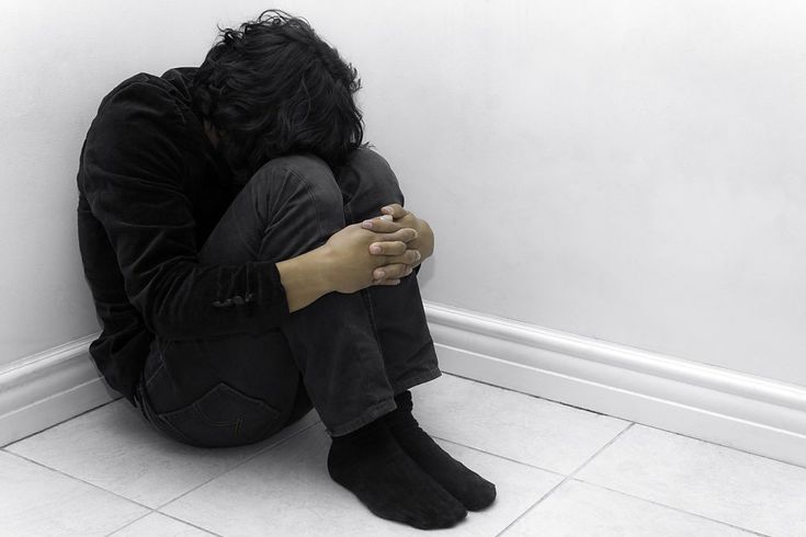 a person is sitting in a corner, looking down at their lap.  they have their arms wrapped around their legs,  giving the impression that they are sad or depressed.