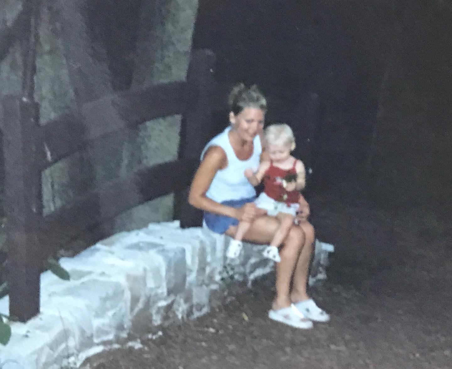 a woman wearing a white tank top and blue shorts is sitting on a stone wall outdoors with a young child on her lap. the child is wearing a red top and white shorts. a wooden fence is behind them. the woman is wearing white slippers.