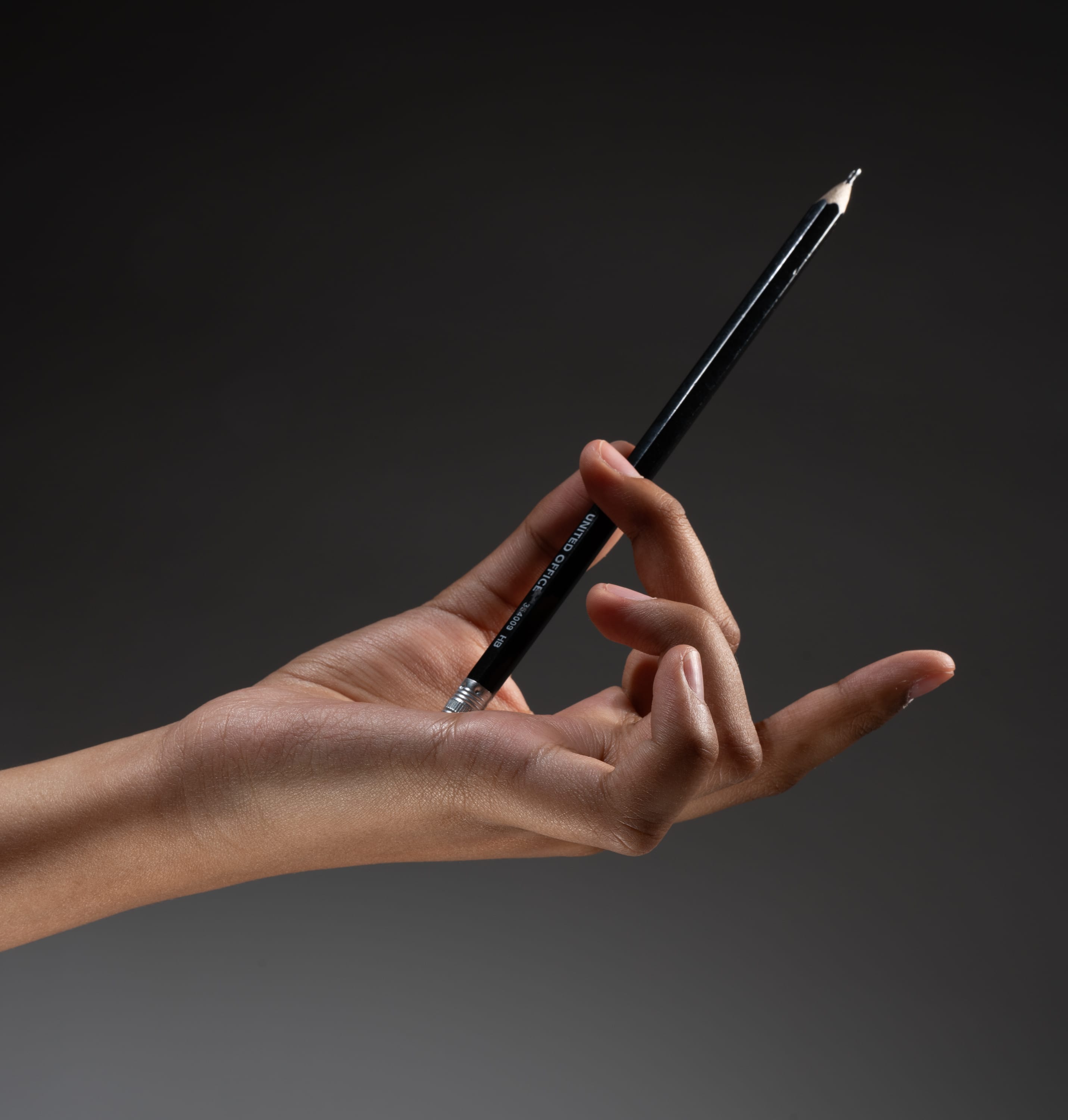 a hand is holding a black graphite pencil against a dark gray background.  the pencil is pointing up and to the right. the hand is illuminated with studio lighting.