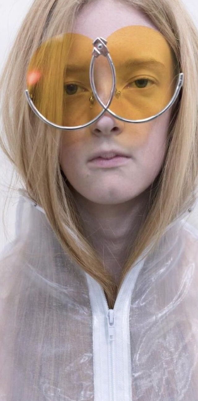 a fairskinned woman with blonde hair is wearing unique yellowtinted sunglasses with a connected silver frame. she is wearing a transparent raincoat zipped up to her neck. the background is a neutral white.