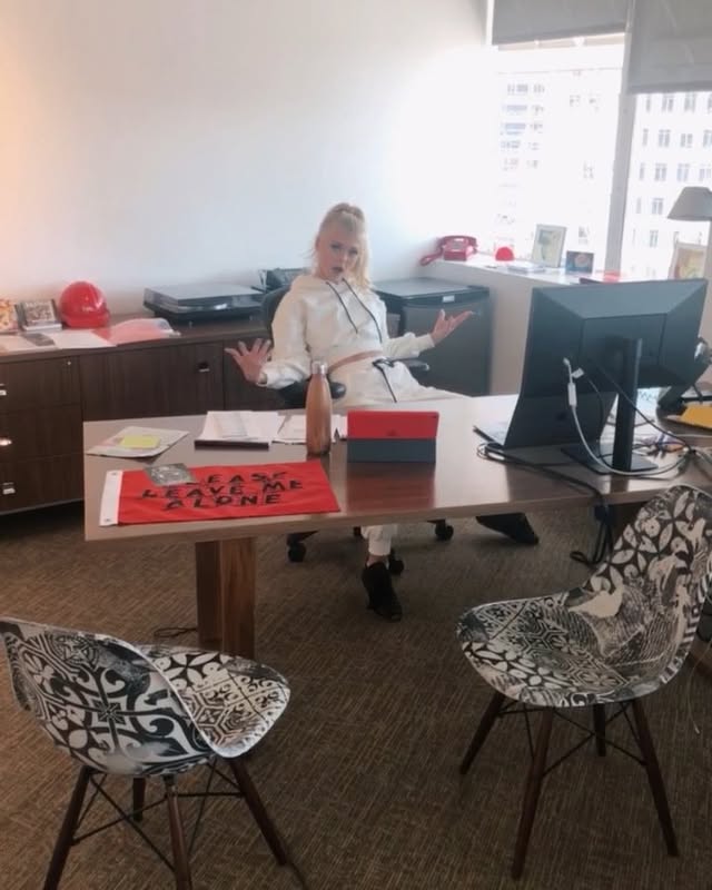 a woman sits at a desk in an office, with her arms outstretched. there is a computer, printer, cabinets, a window, a red hard hat, a water bottle, a red box, and a "leave me alone" sign on the desk. there are two chairs in front of the desk.