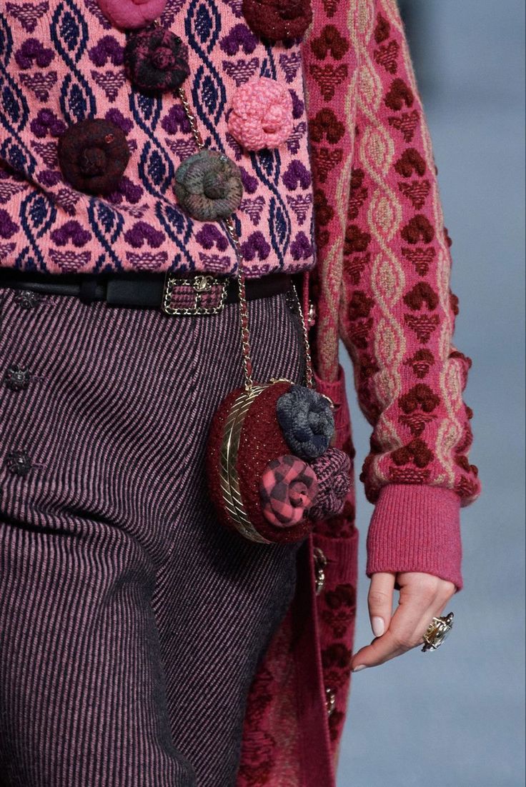 closeup view of a woman's outfit, focusing on a richly textured pink and purple knit cardigan and matching pinstriped trousers. the cardigan is intricately detailed with various textured embellishments and flowershaped appliqués.  a matching maroon handbag with similar floral detailing hangs from a chain across her body.  a gold ring is visible on one of her fingers. the overall aesthetic is highfashion and autumnal in color palette.  the image suggests a runway or photoshoot setting.