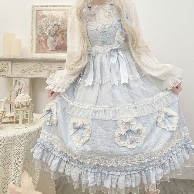 a woman is wearing a long blue and white lolita style dress. the dress is adorned with lace and heartshaped decorations. the background is a white room with a painting on the wall. the photo is taken from eye level and uses natural lighting.