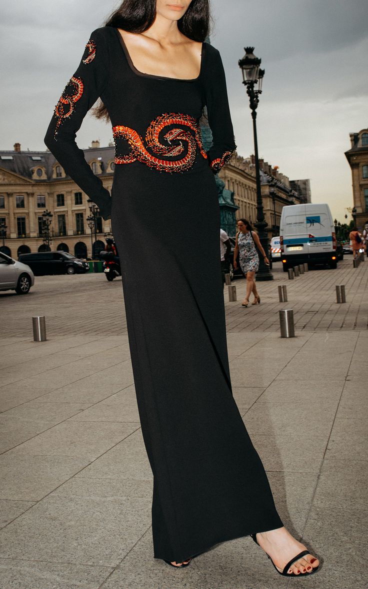 a woman stands on a paved area in a city, wearing a longsleeved black dress with a square neckline and decorative embellishments. the dress features spiral designs in orange beads on the sleeves and around the waist. she is wearing black sandals with thin straps. the background includes a street scene with buildings, a lamppost, a bus, pedestrians, traffic cones, and barriers.